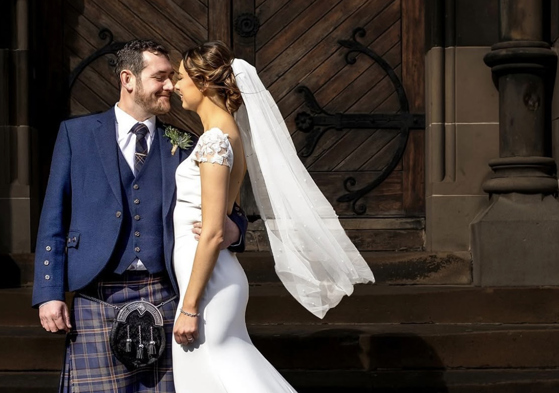 Bride with veil and groom wearing a kilt standing outside venue in an embrace while smiling at each other